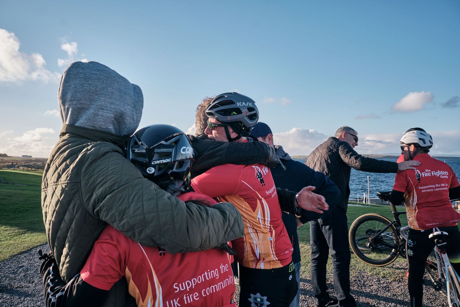 Cyclists in helmets share a group hug outdoors, wearing jerseys supporting UK fire communities on a cloudy day.