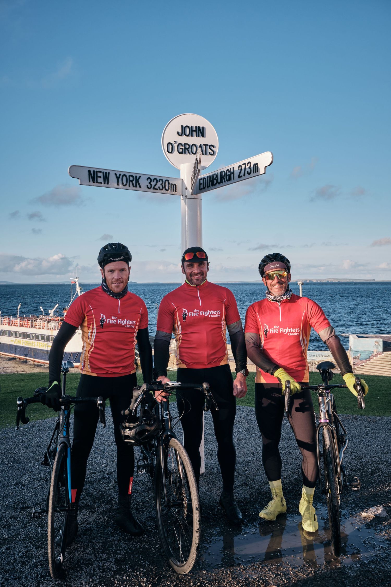 Three cyclists in red jerseys pose with bikes at John O'Groats signpost showing New York and Edinburgh distances.