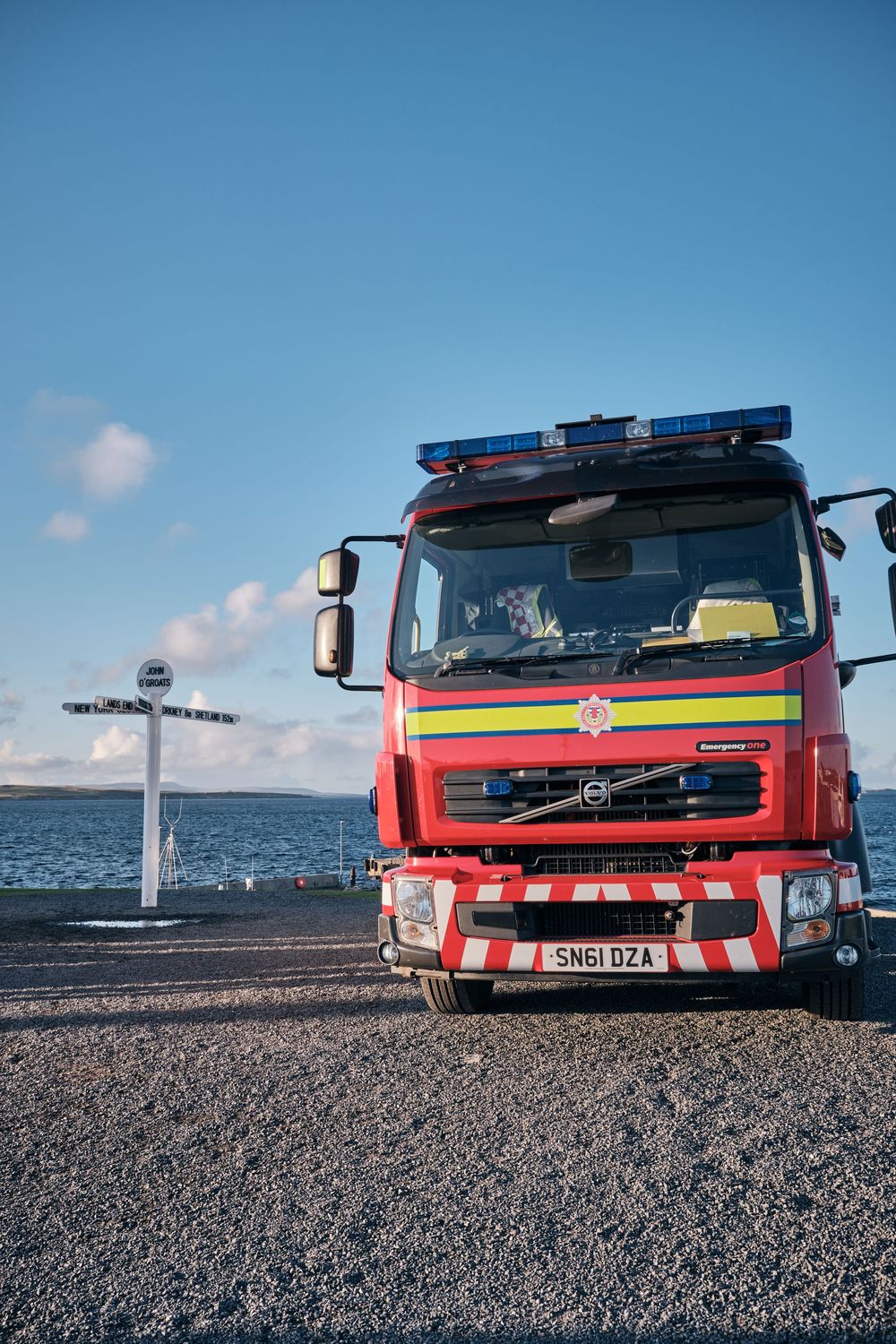 Red Volvo fire engine truck with blue lights parked near waterfront on gravel, registration SN61 DZA, Scotland.