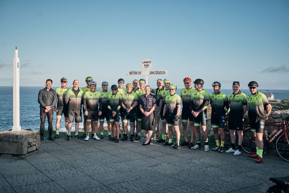 Group of cyclists in green jerseys posing together on a seaside promenade under a clear blue sky.