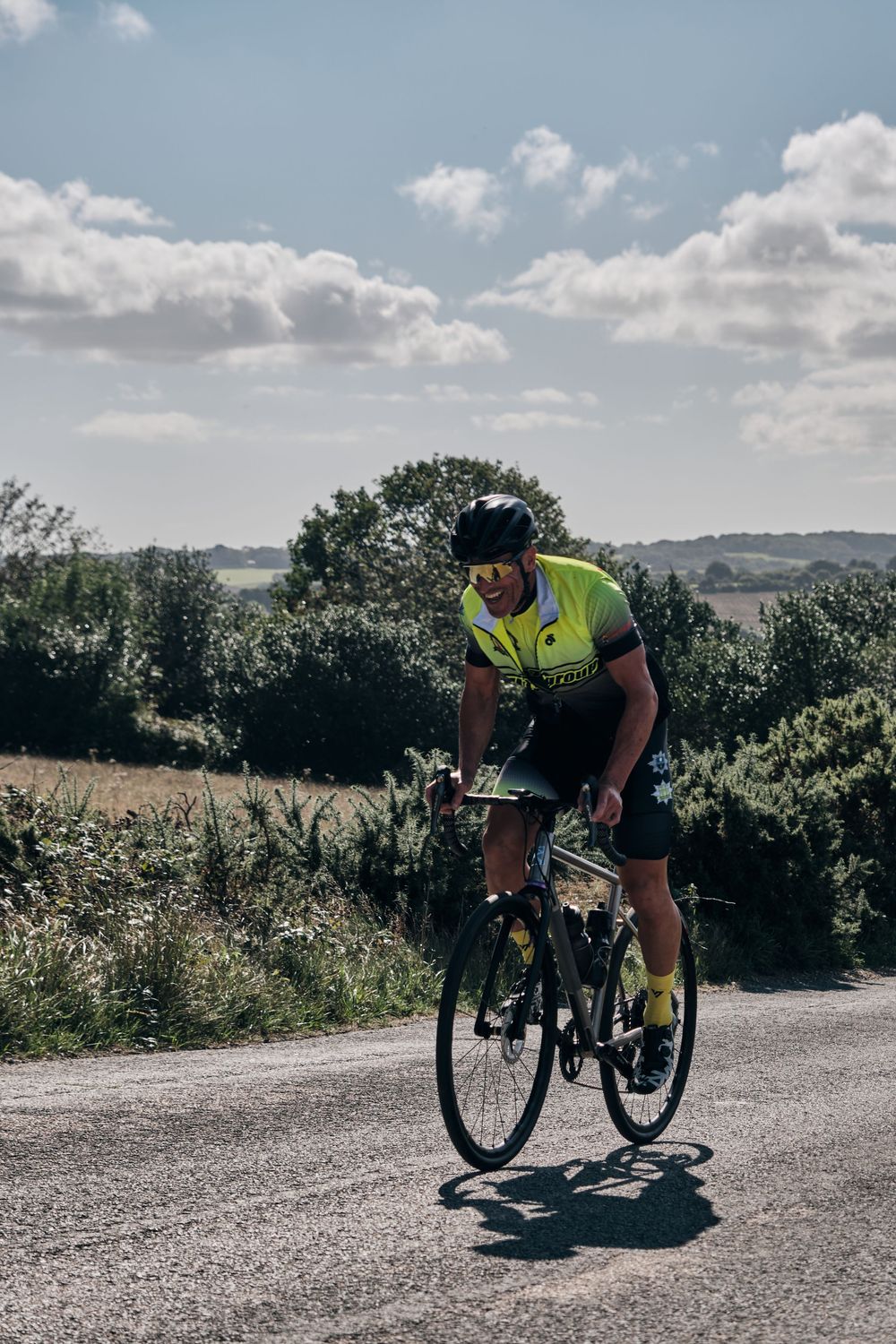Cyclist in neon yellow jersey riding a road bike on a sunny rural road surrounded by green trees.