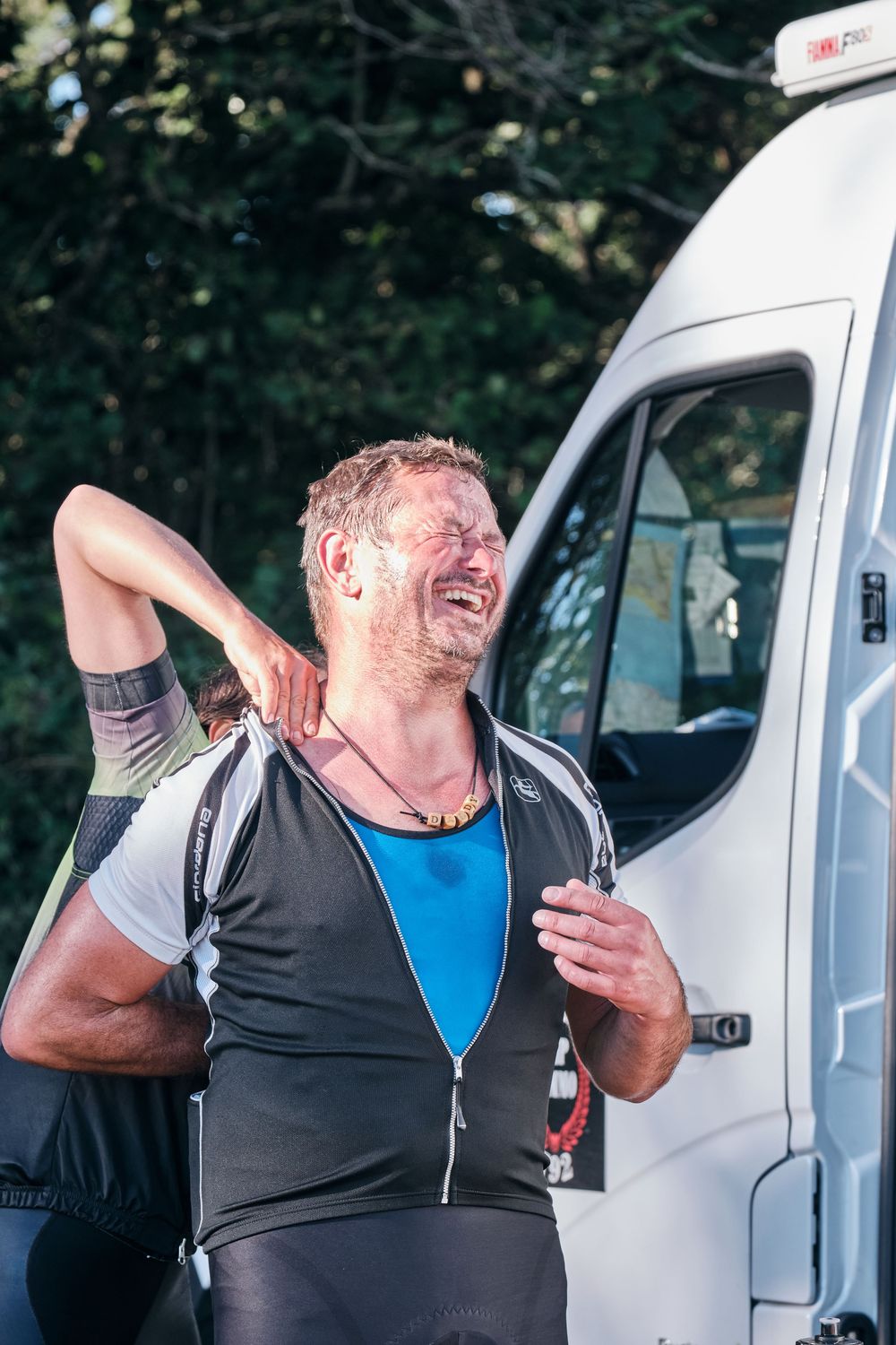 Male cyclist in black and blue jersey laughing beside a white van after a race, with trees in background.