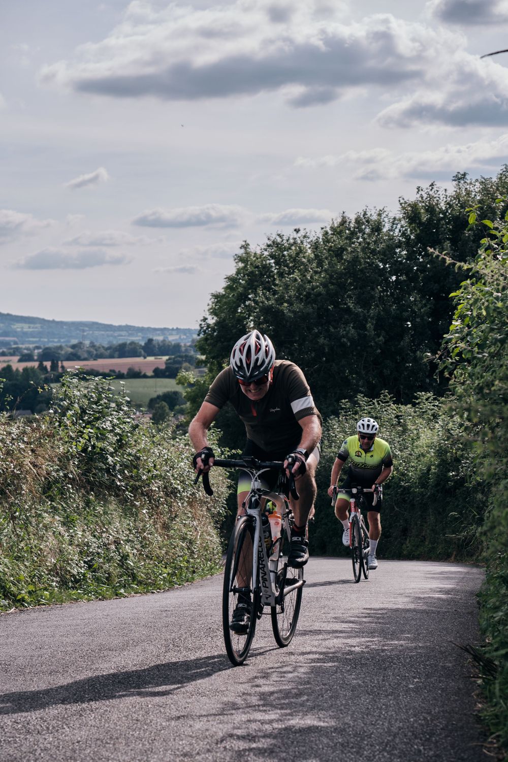 Cyclists in helmets ride road bikes down a narrow country lane surrounded by lush green hedgerows and fields.