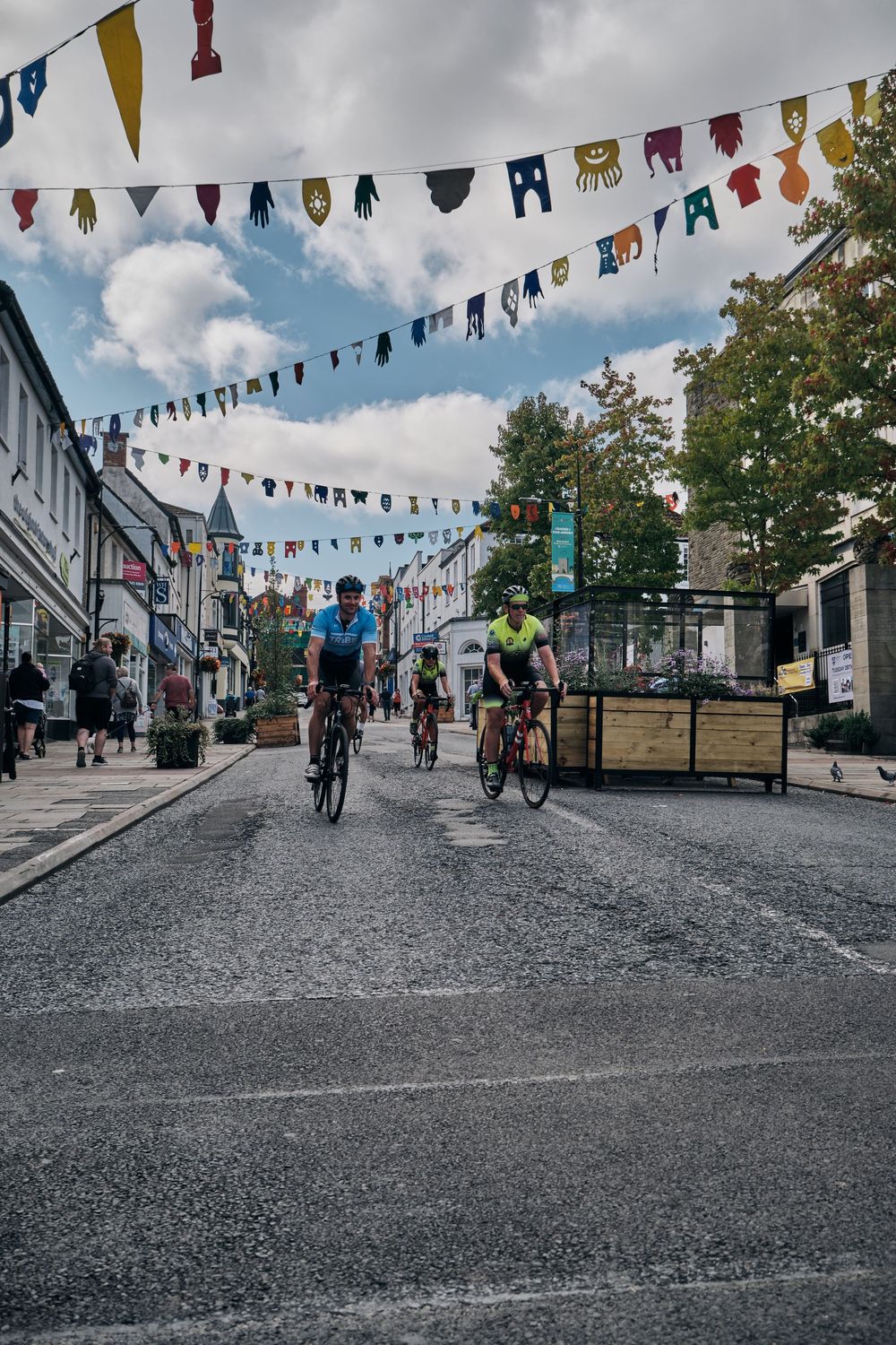Cyclists riding through a festive street decorated with colorful bunting flags on a partly cloudy day.