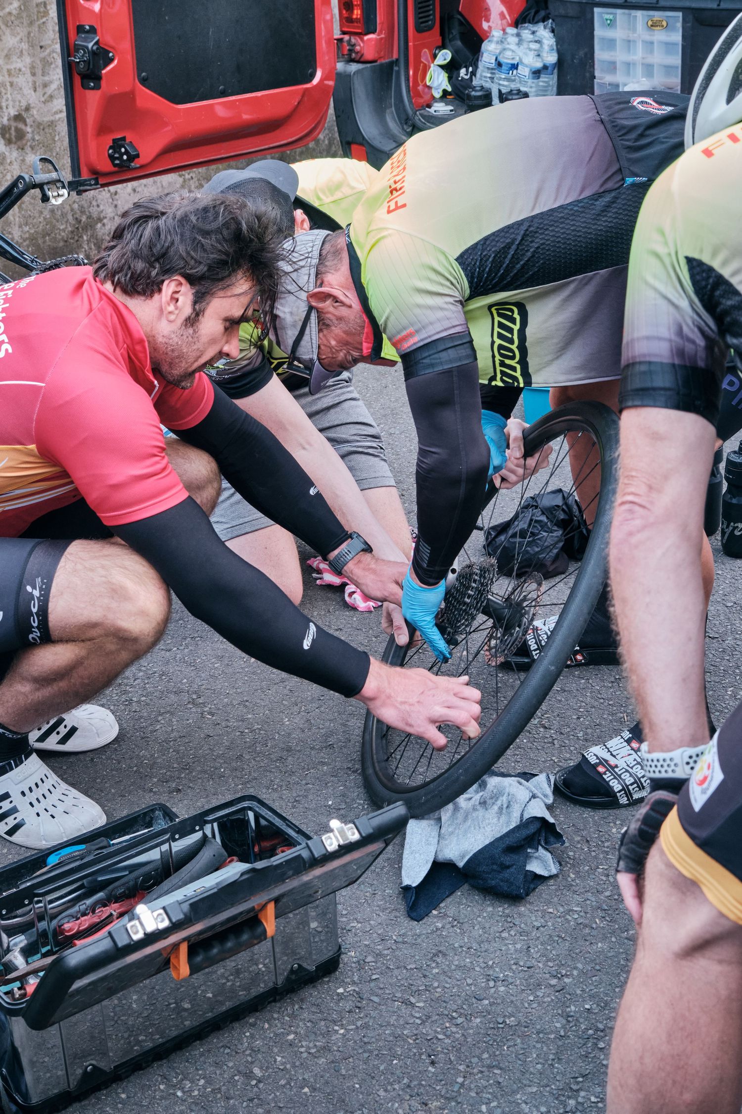 Cyclists repairing a bike wheel roadside with tools and blue tape during a cycling race or event.