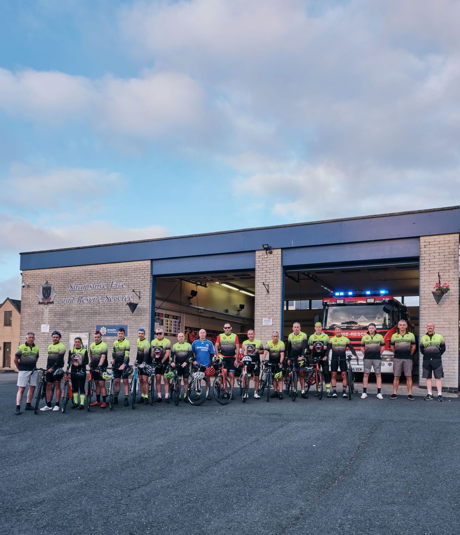 Large group of cyclists in matching jerseys posing outside a fire station before a charity ride.
