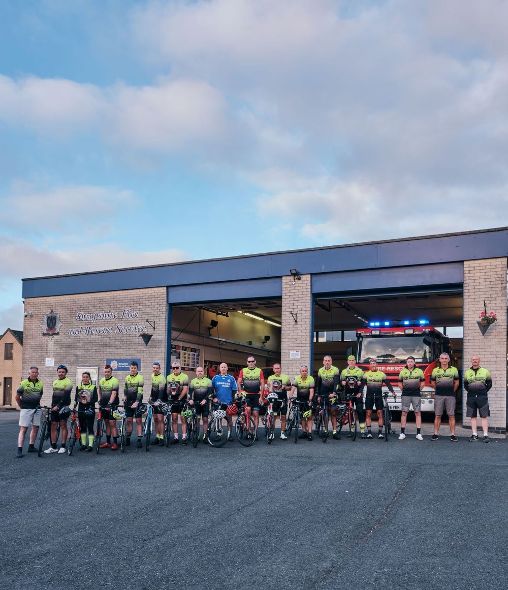 Large group of cyclists in matching jerseys posing outside a fire station before a charity ride.