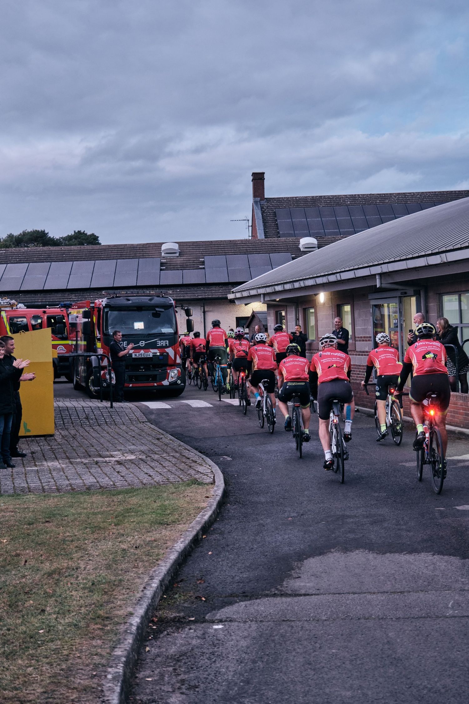 Cyclists in red jerseys riding through a parking area near a building on a cloudy day.