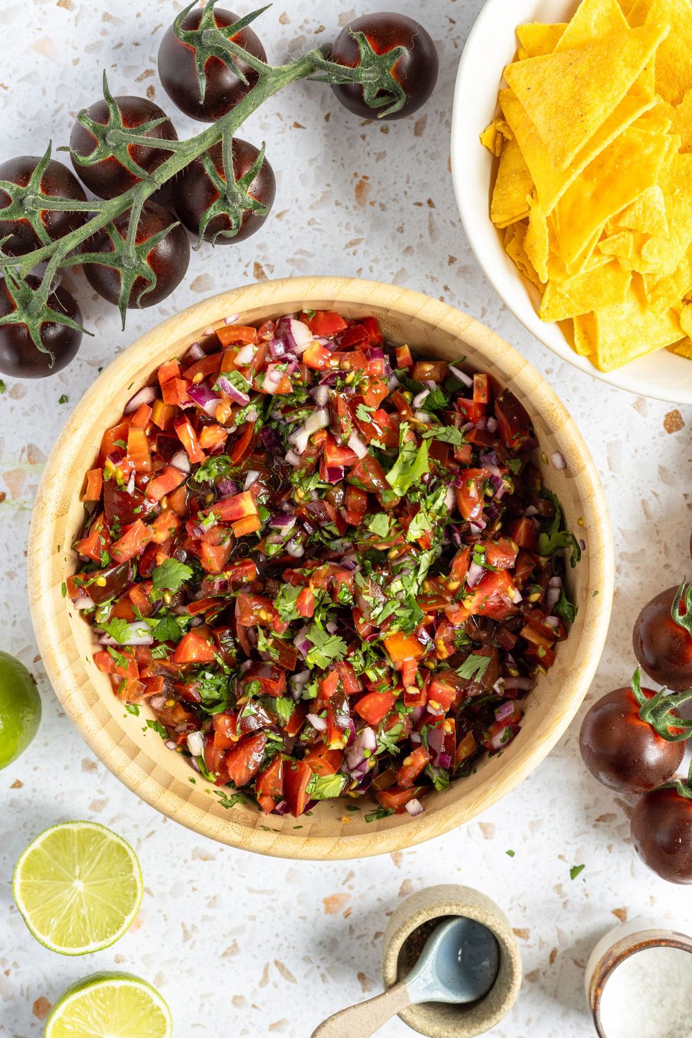 Fresh homemade salsa in wooden bowl with cherry tomatoes, lime, tortilla chips, and cilantro on white surface.