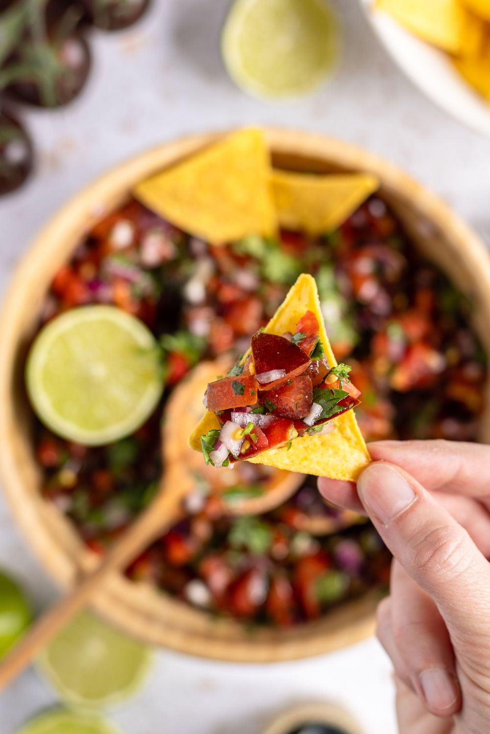Hand holding a tortilla chip topped with fresh pico de gallo salsa over a wooden bowl of black bean salsa.