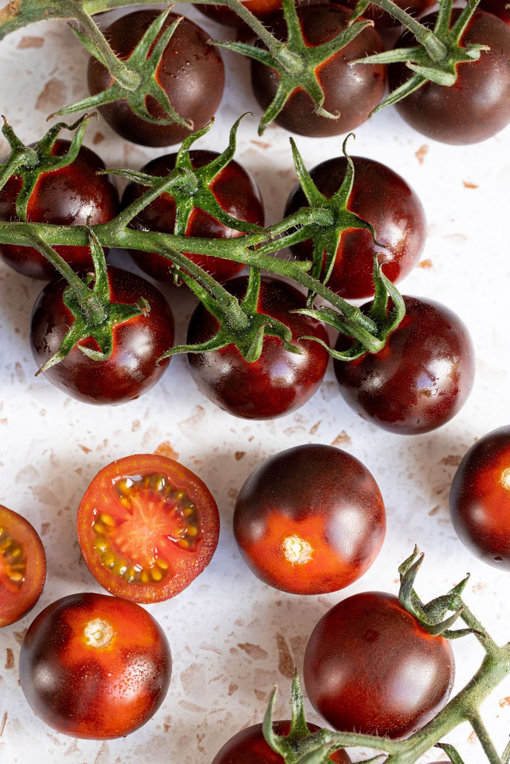 Dark purple cherry tomatoes on the vine, some halved to reveal red interior, displayed on a white surface.