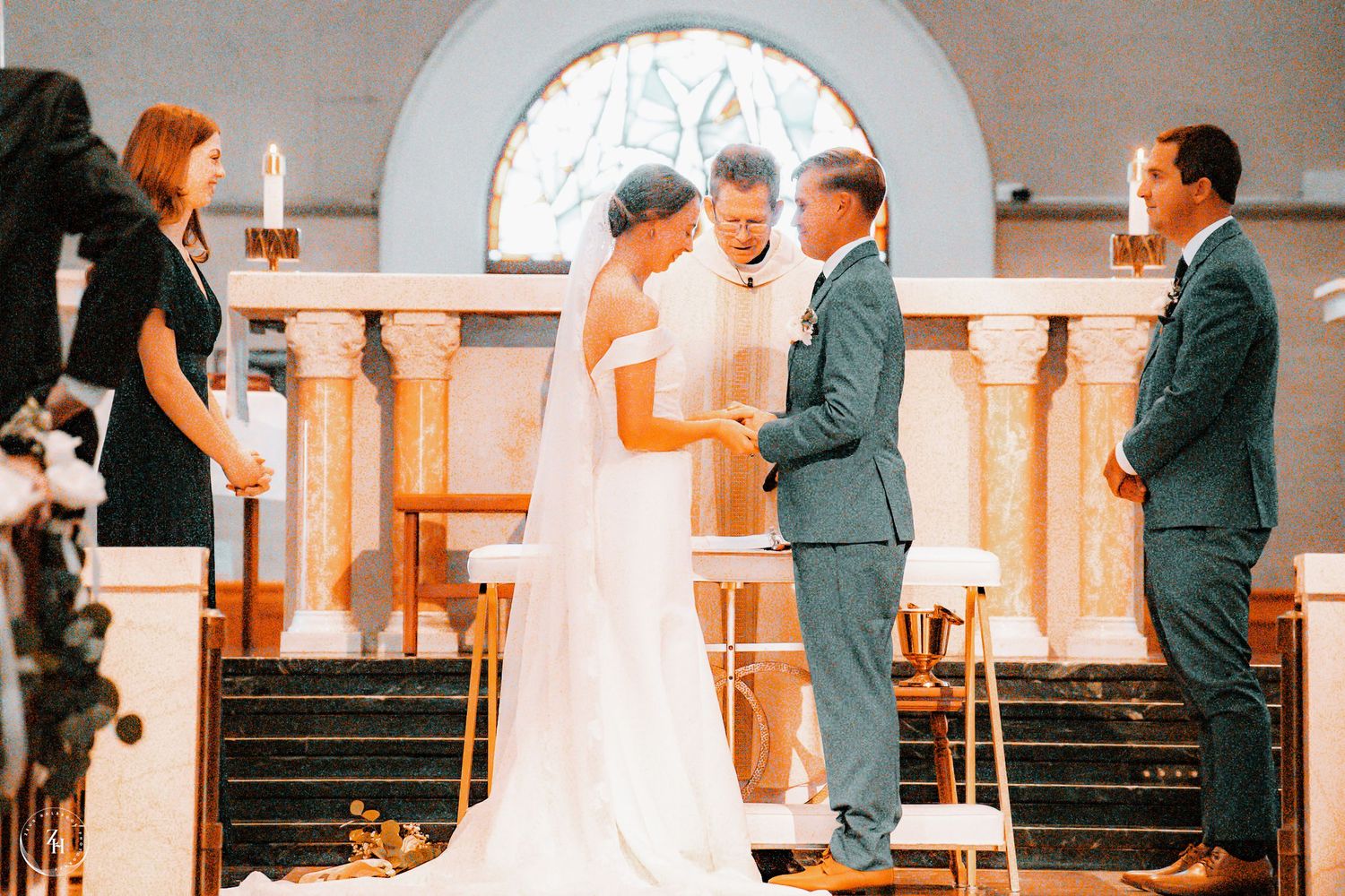 Bride and groom exchange vows at church altar with officiant and wedding party during elegant indoor ceremony.