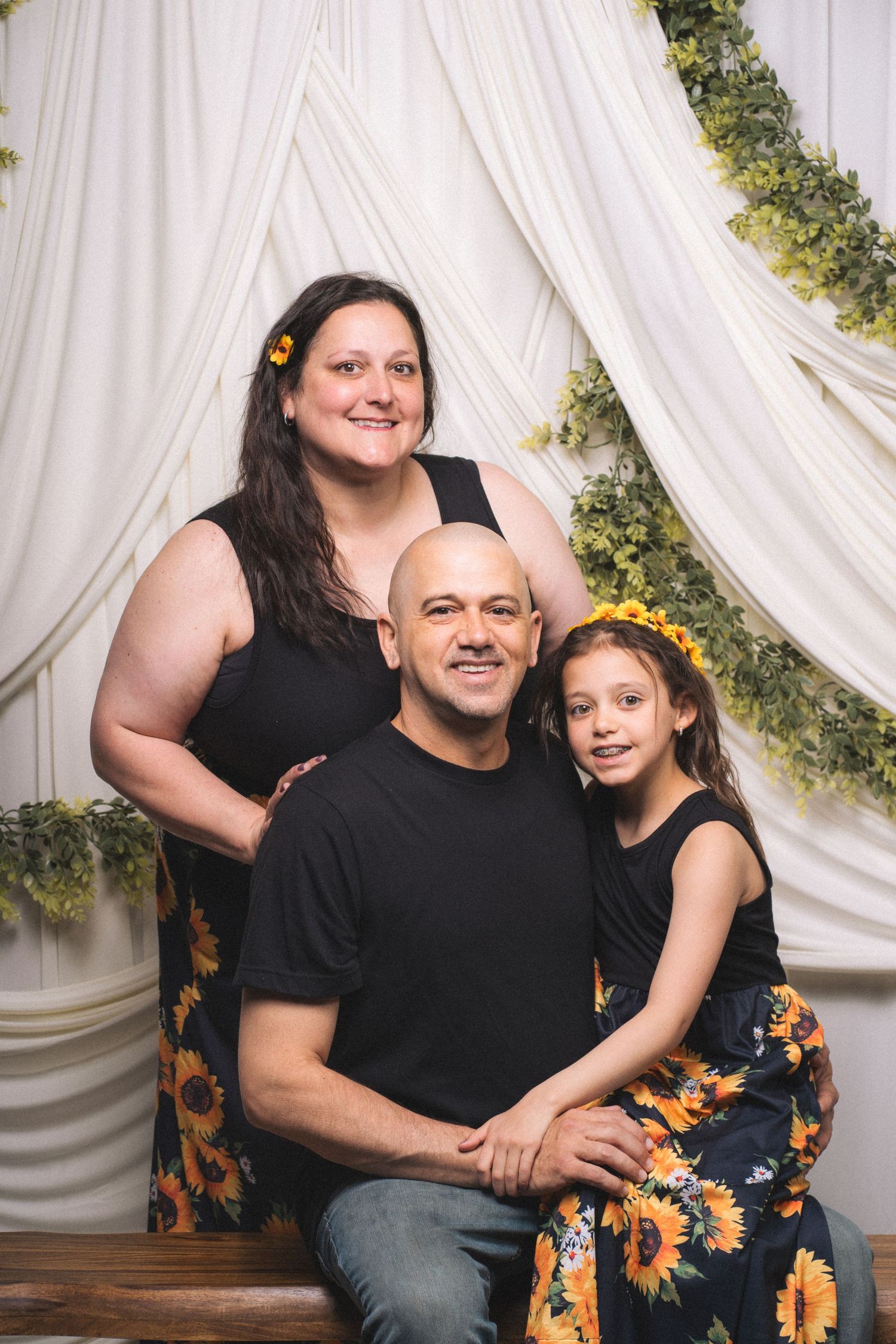 Family of three posing for a portrait with white drape backdrop and greenery, wearing black and sunflower outfits.