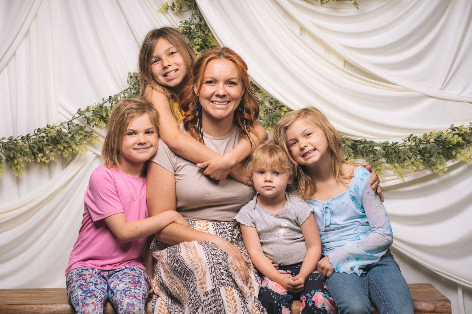 A smiling redhead woman poses with four young girls in front of a white draped backdrop with greenery.