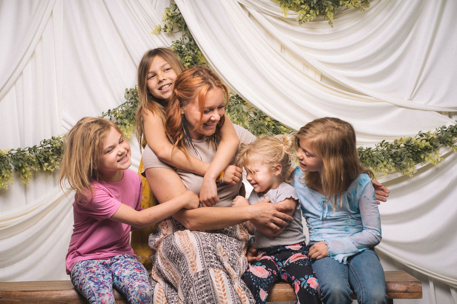 A joyful woman and four young girls laugh together under a floral canopy in a bright, cheerful photo session.