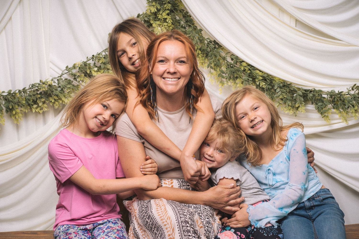 A smiling redhead woman poses with four young children in a bright studio with greenery backdrop.