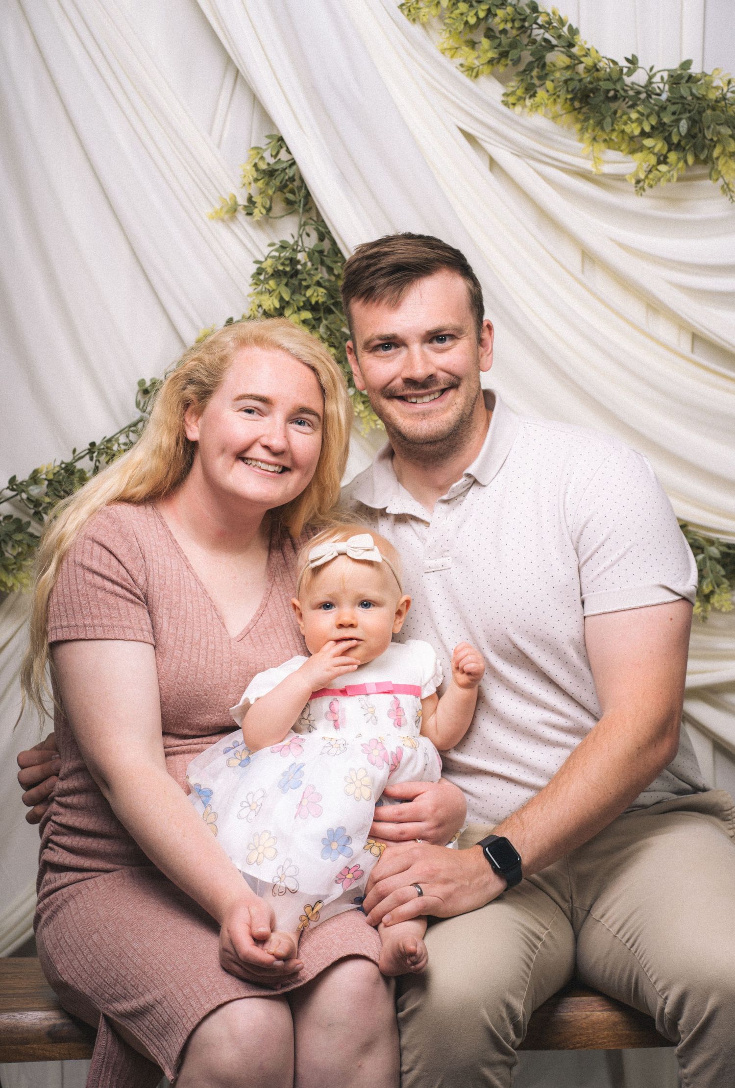 Happy young family portrait with smiling couple and baby girl in floral dress, posed before elegant white draped backdrop.