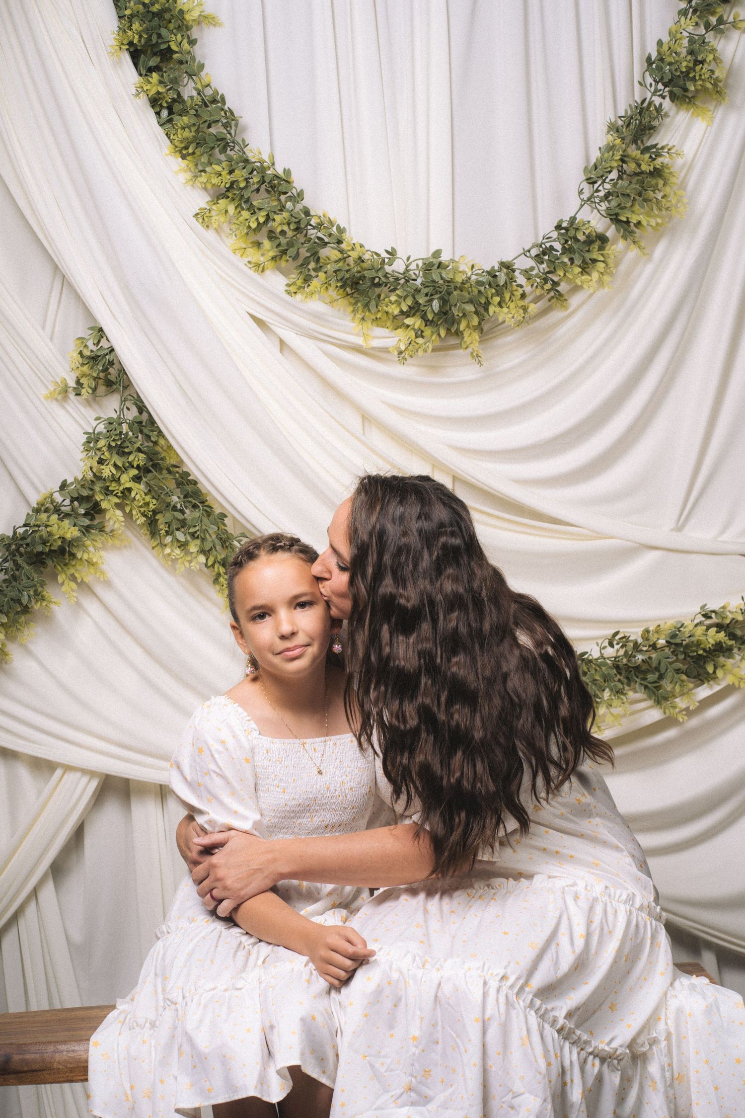 A mother in white dress kisses her young daughter on the cheek against elegant white drape backdrop with green garlands.
