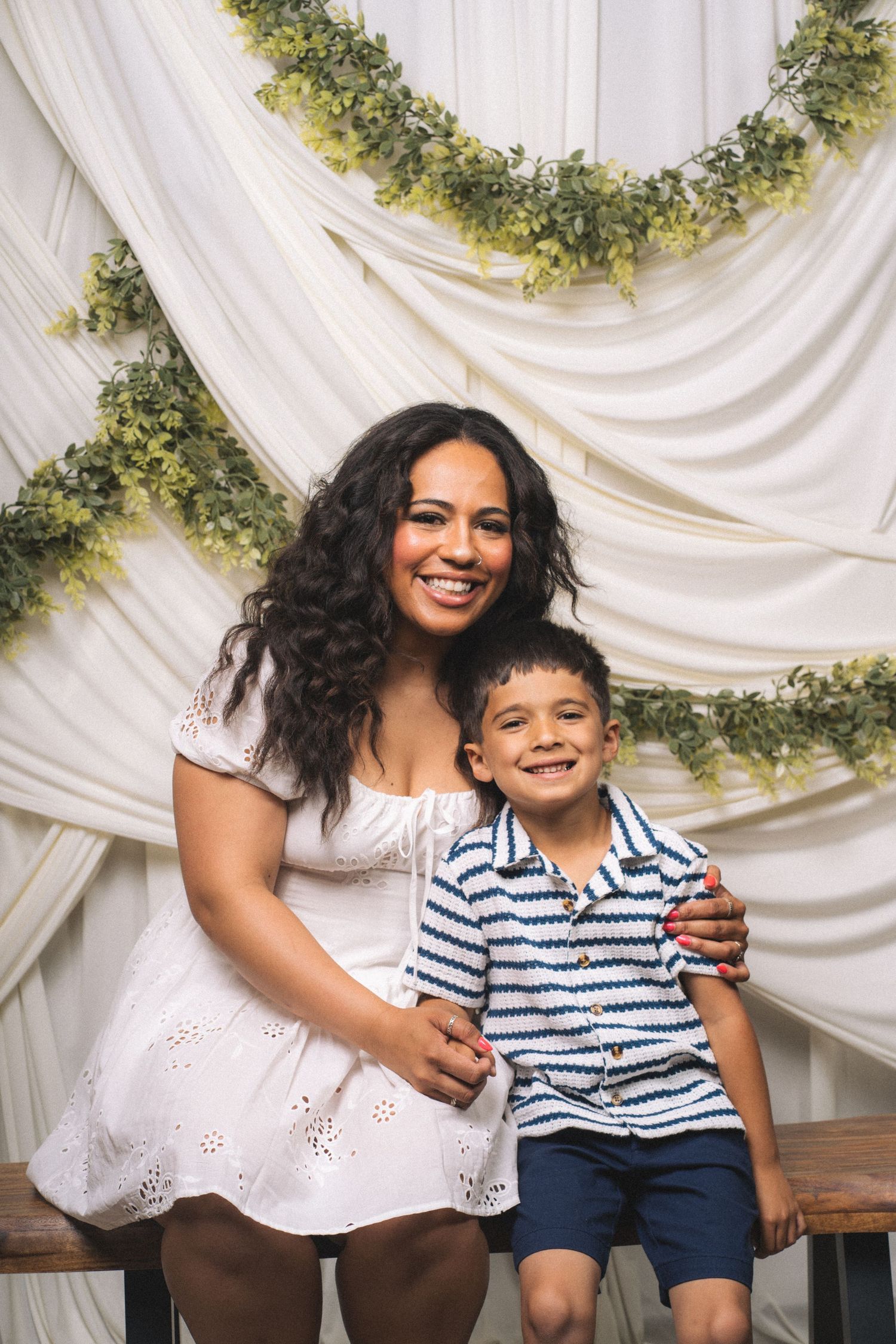 A smiling woman in a white dress poses with a young boy in a striped shirt against a white draped backdrop with greenery.