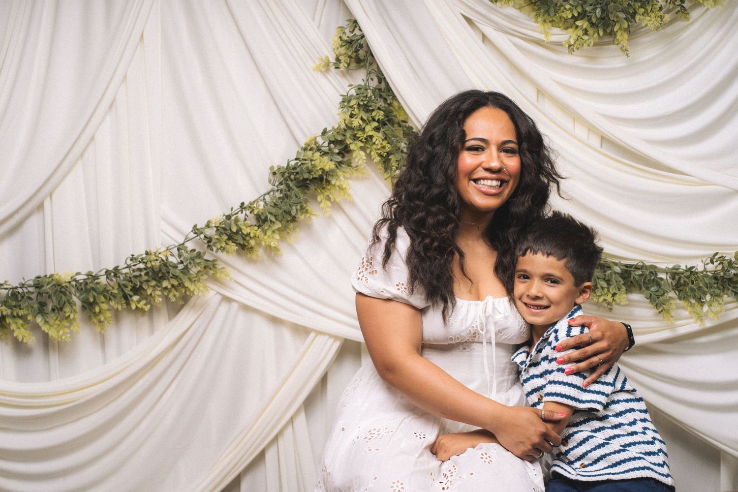 Woman in white dress smiling with young boy in striped shirt, posing in front of elegant white draped backdrop.