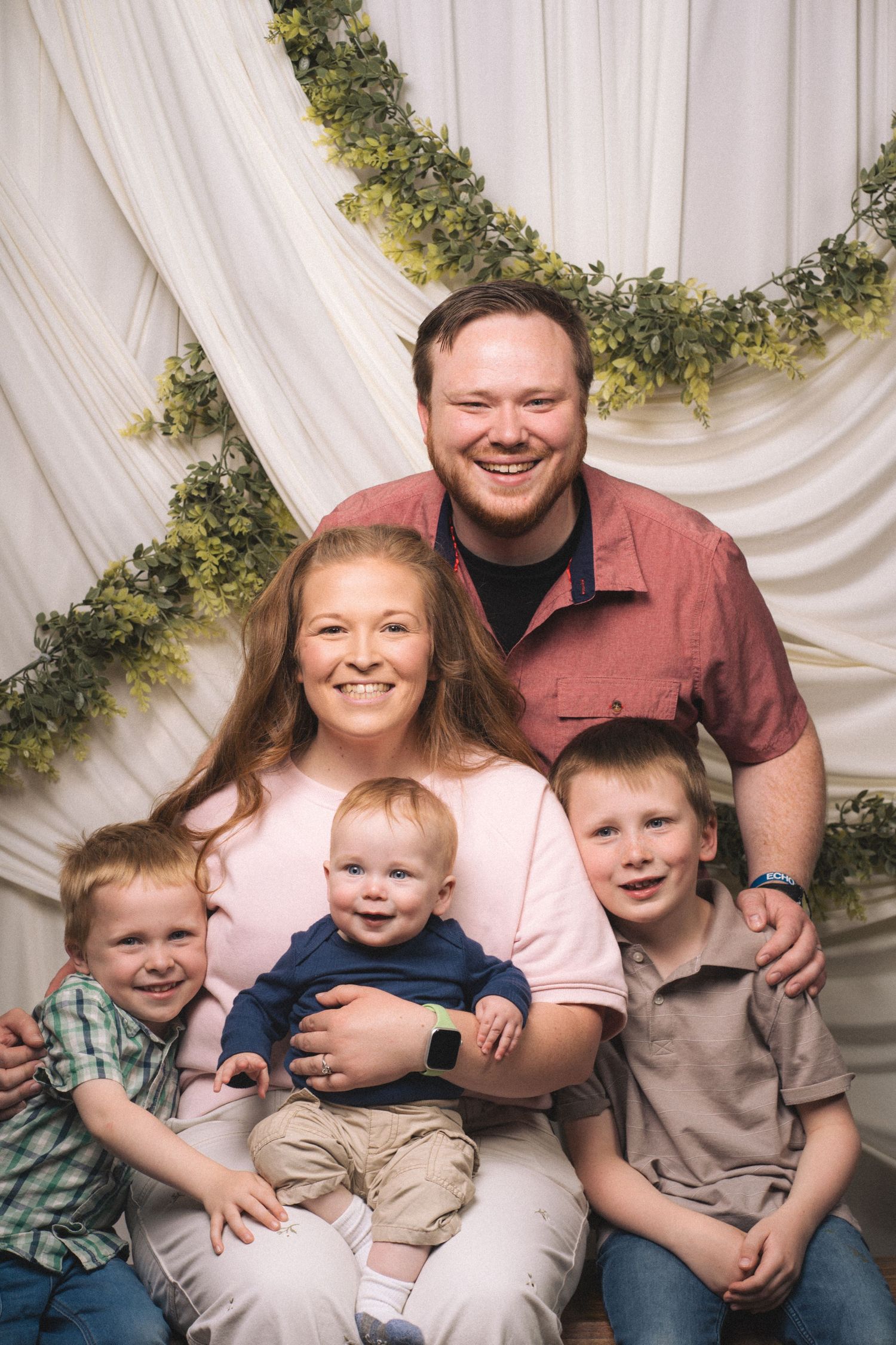 A happy family of five poses for a portrait with green garland backdrop, including parents and three young boys.