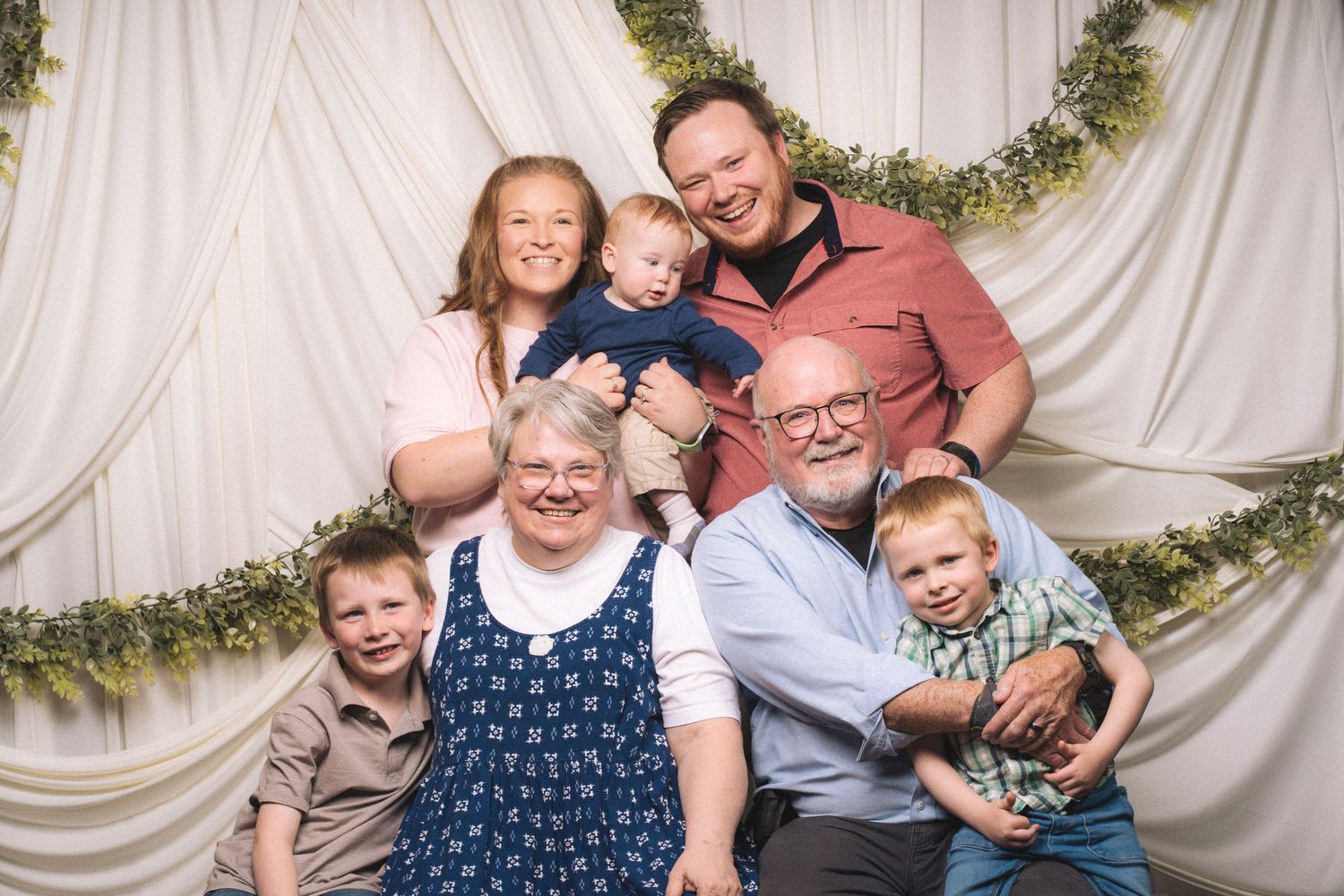 Multi-generational family portrait with grandparents, parents, and three young children posing together in front of white draped backdrop.
