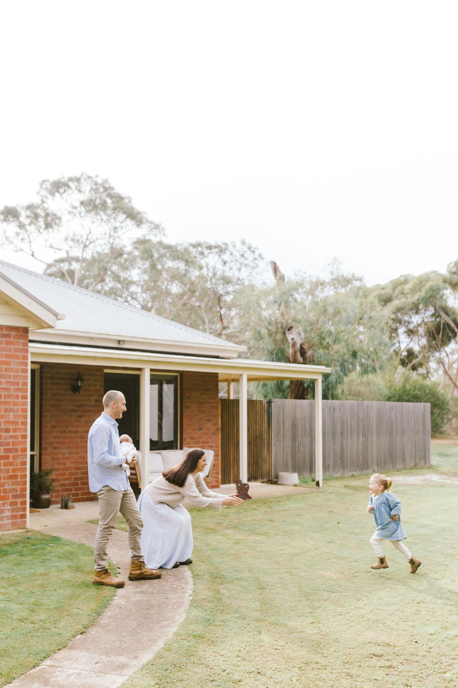 Family playing in backyard of brick home with lush green lawn, surrounded by eucalyptus trees on a bright day.