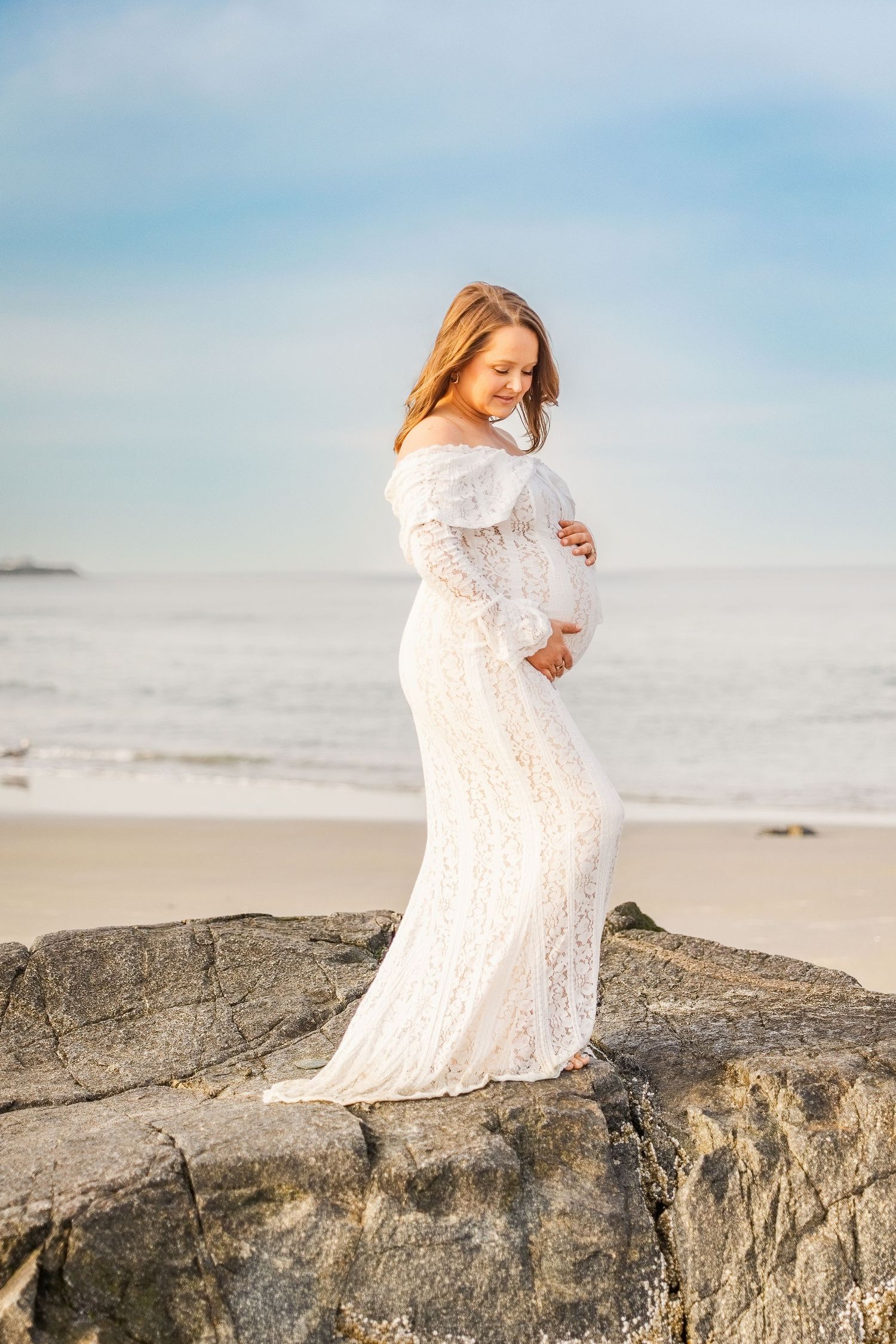 Pregnant woman in white lace gown standing on beach rocks, cradling her bump, with ocean waves in the background.