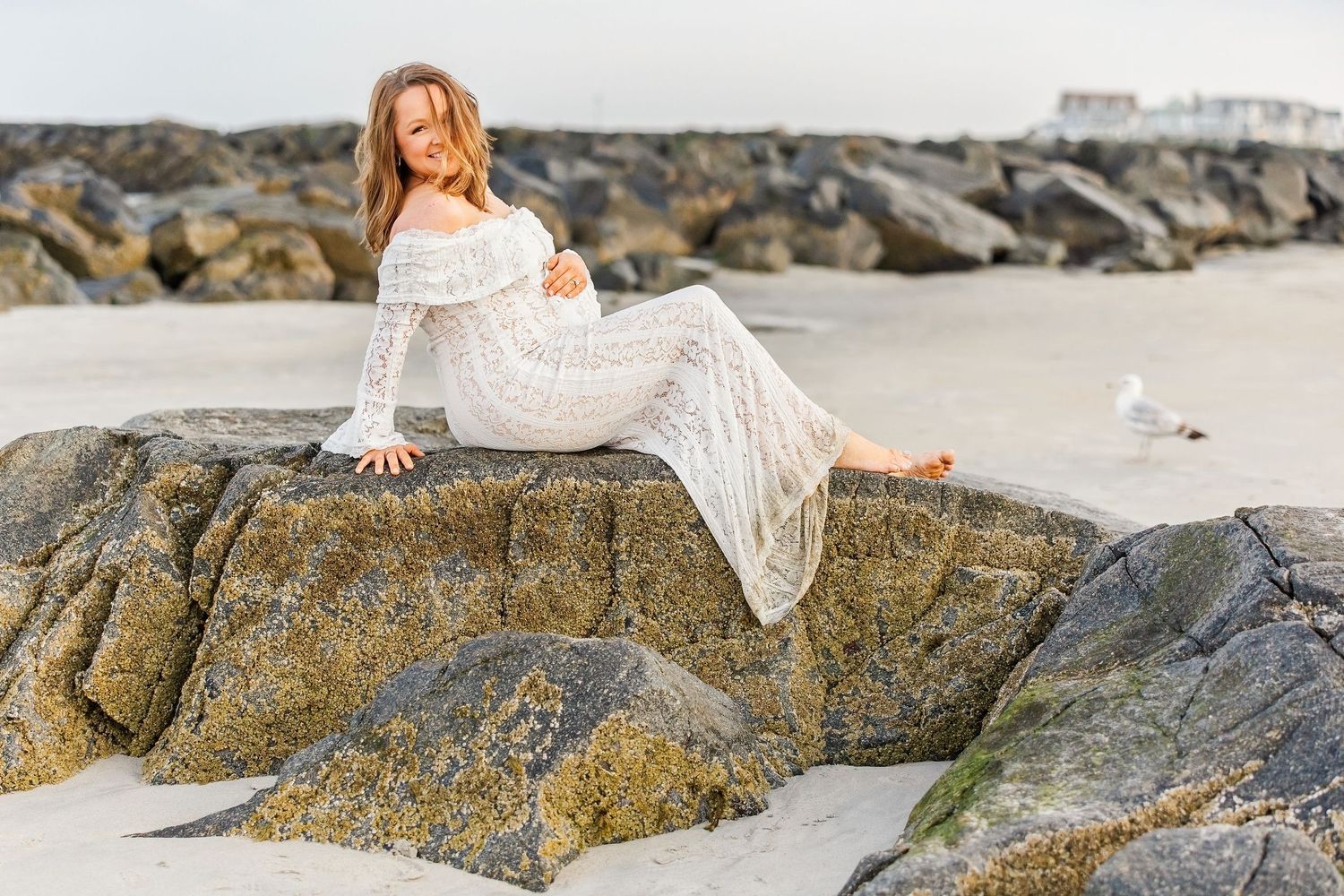 A woman in a white lace dress sits gracefully on coastal rocks at a sandy beach with a seagull nearby.