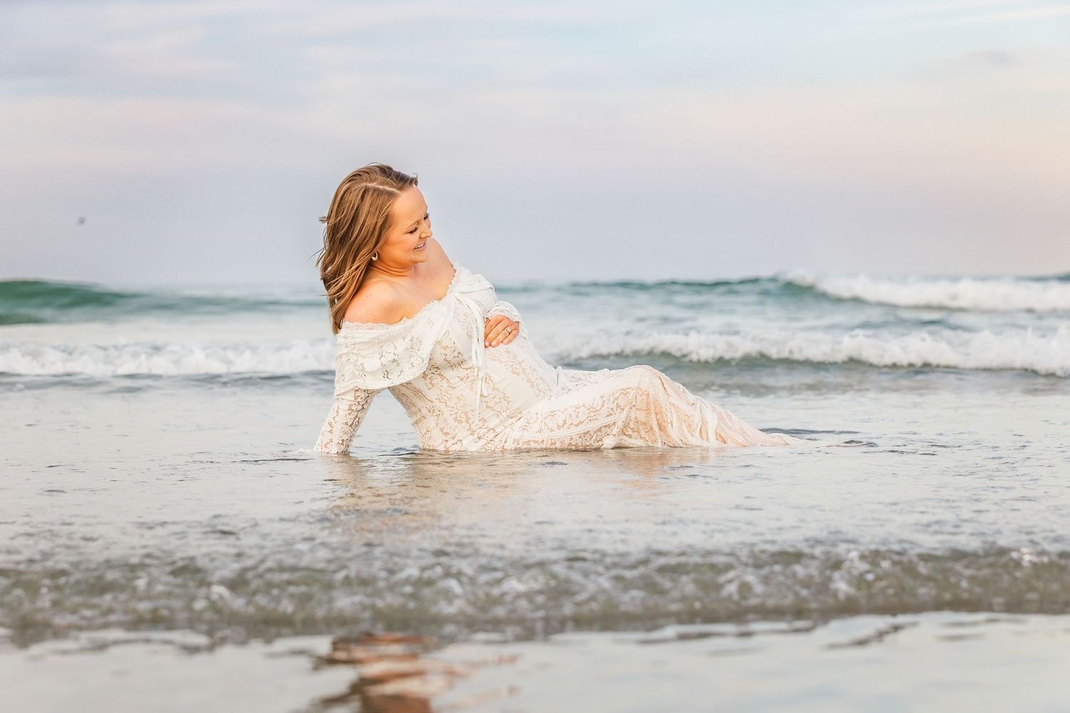 Pregnant woman in white lace dress posing gracefully in ocean waves on a beach during golden hour.