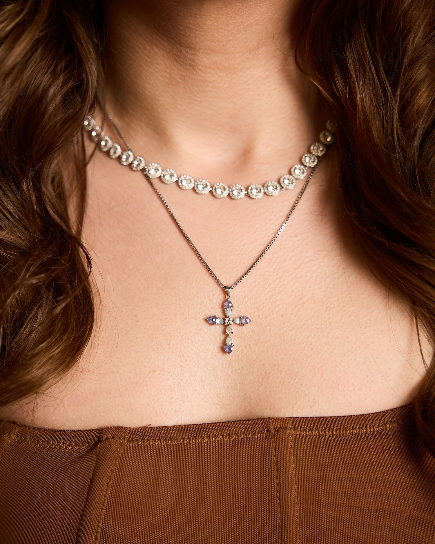 Woman wearing layered necklaces featuring a crystal tennis choker and a gemstone cross pendant on a delicate chain.