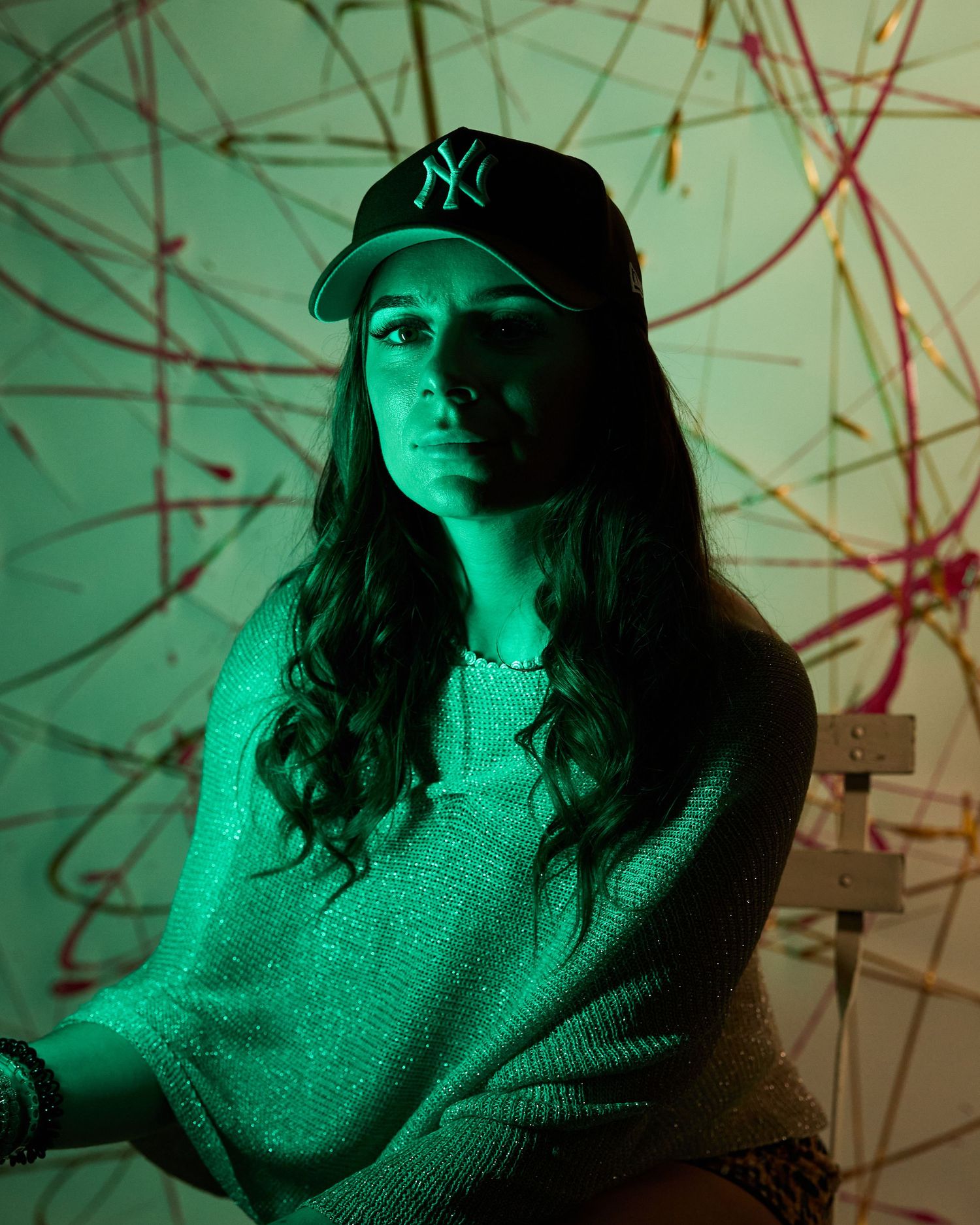 Woman in NY Yankees cap and green sweater posed under green lighting with abstract line art background.