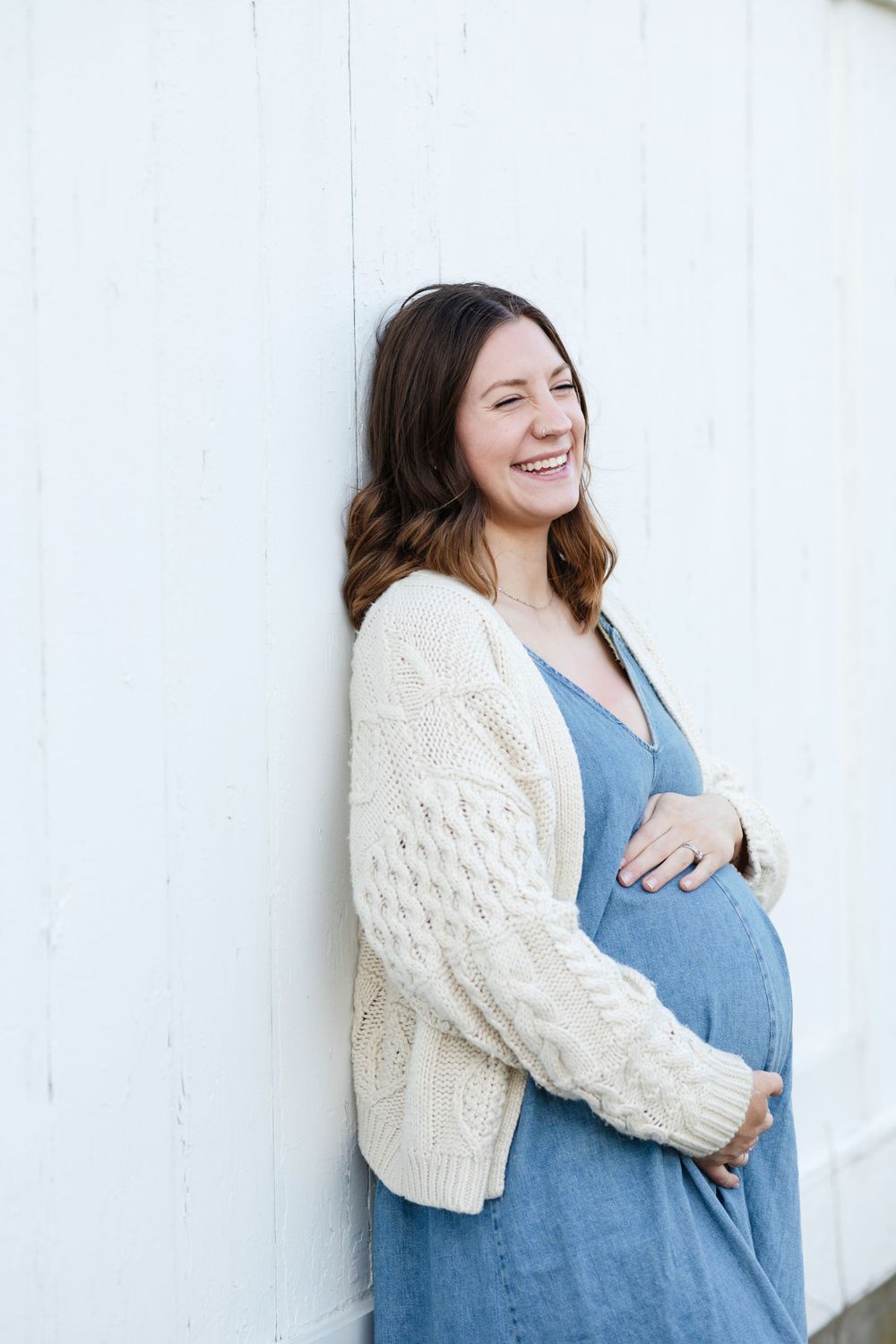 Laughing expectant mother wears a jean dress and chunky sweater leaning on white barn for maternity photos. Andover maternity photographer