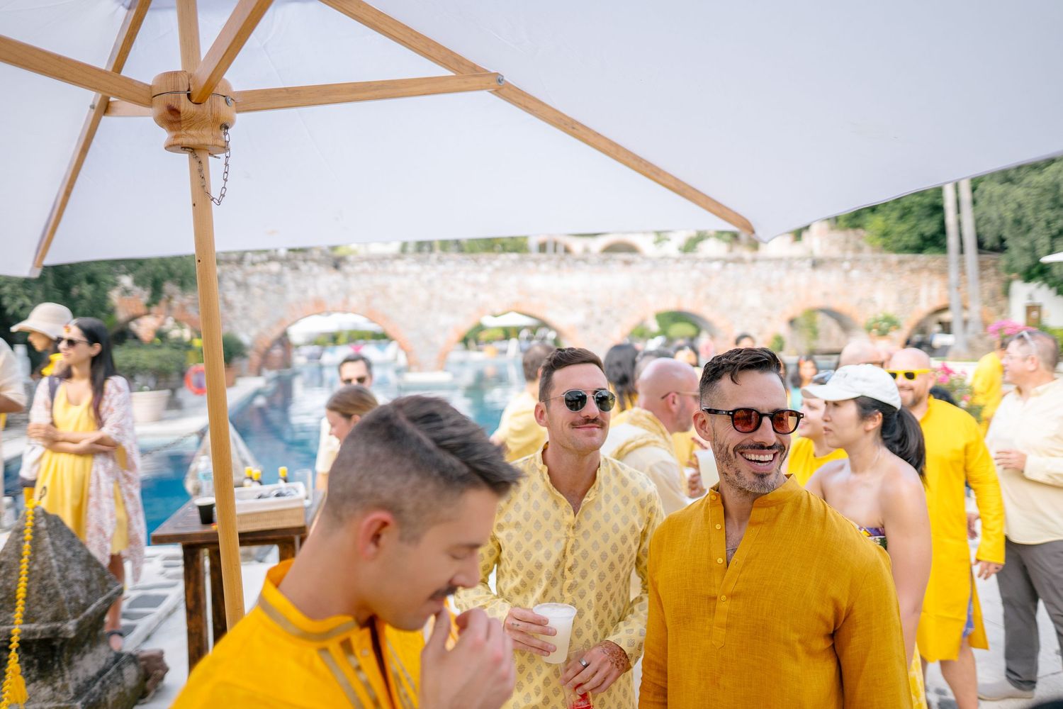 Men in matching yellow kurtas laughing and socializing at an outdoor poolside event on a sunny day.