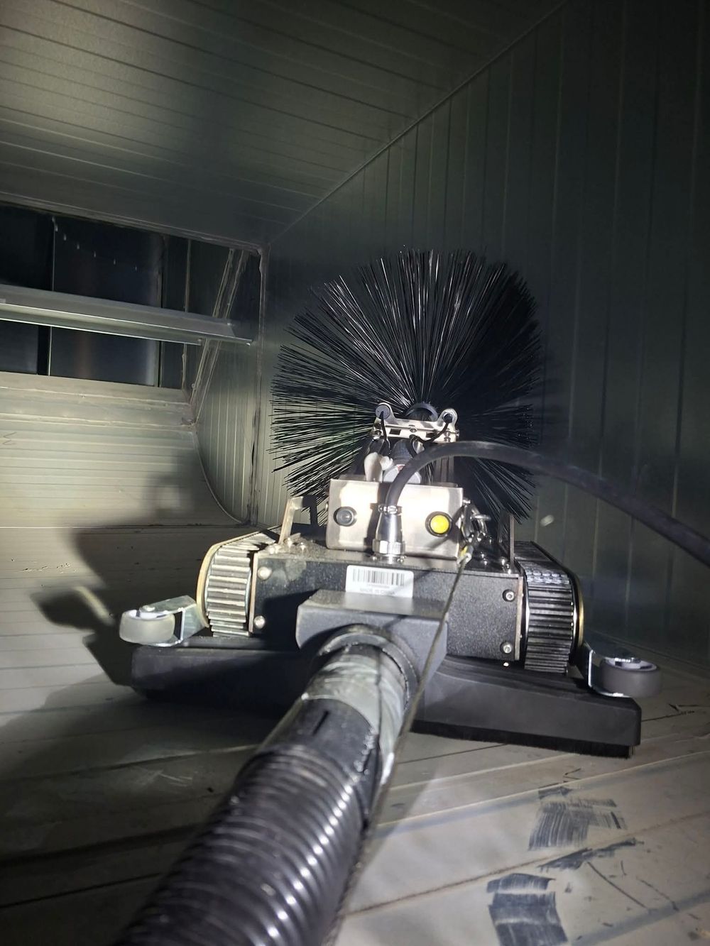 Air duct cleaning robot with rotary brush and vacuum hose inside a large HVAC duct system.