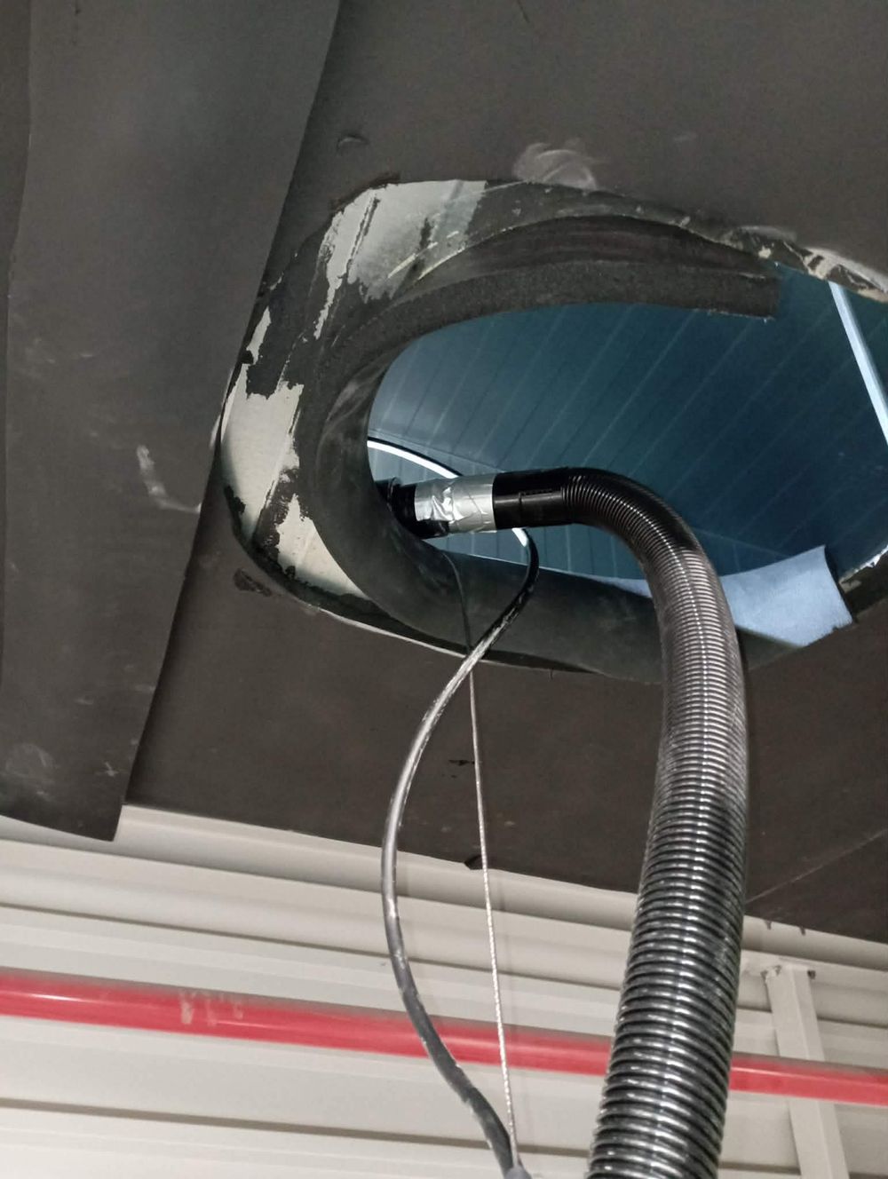 A flexible corrugated hose inserted into a ceiling duct opening with visible wiring and foam insulation around the edges.