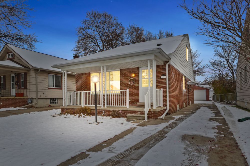 Brick bungalow with white porch railings and snowy front yard at dusk, featuring a detached garage and mature trees.