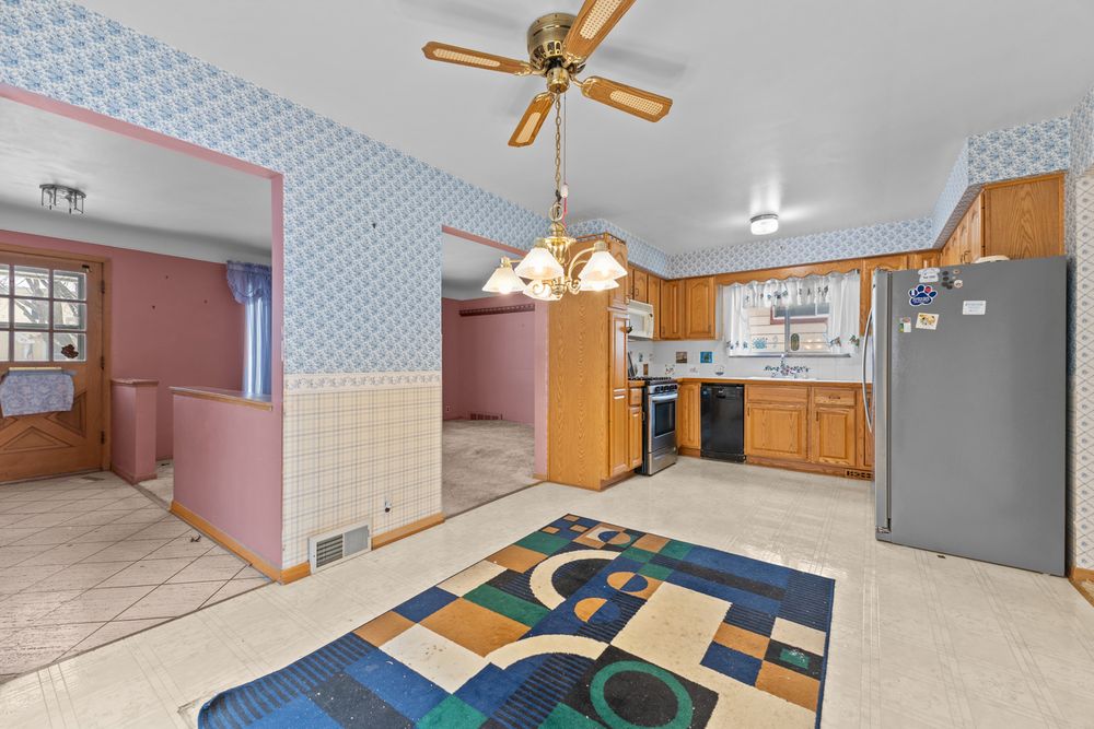 Kitchen with ceiling fan, wood cabinets, colorful area rug, and blue wallpaper adjoining a pink dining area.