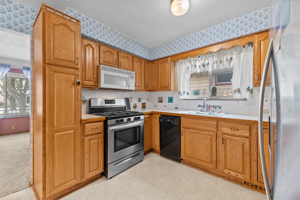 Oak kitchen with stainless gas range, microwave, black dishwasher, and fridge, featuring blue wallpaper and tile backsplash.