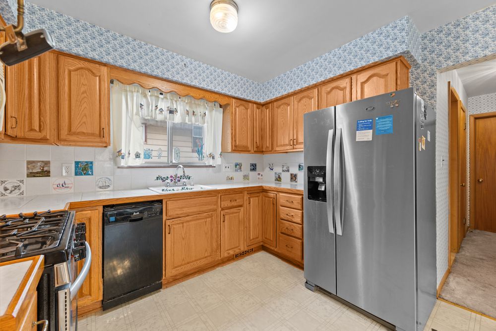 Spacious kitchen with oak cabinets, stainless steel fridge, black dishwasher, gas range, and blue floral wallpaper.