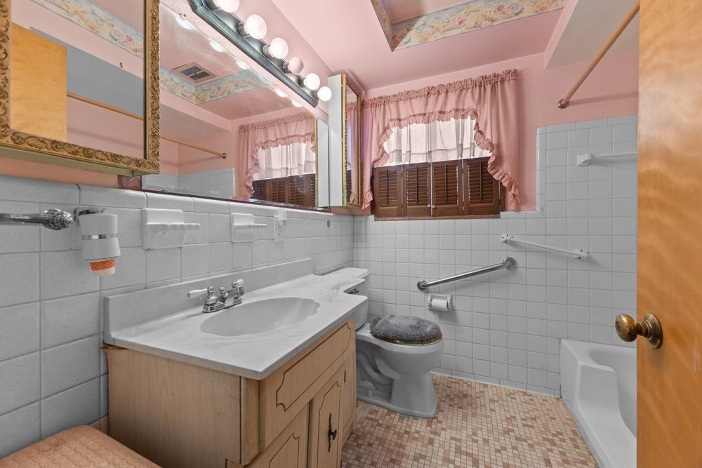 Pink-walled bathroom with white tile, vanity sink, toilet, and bathtub featuring grab bars and curtained window.