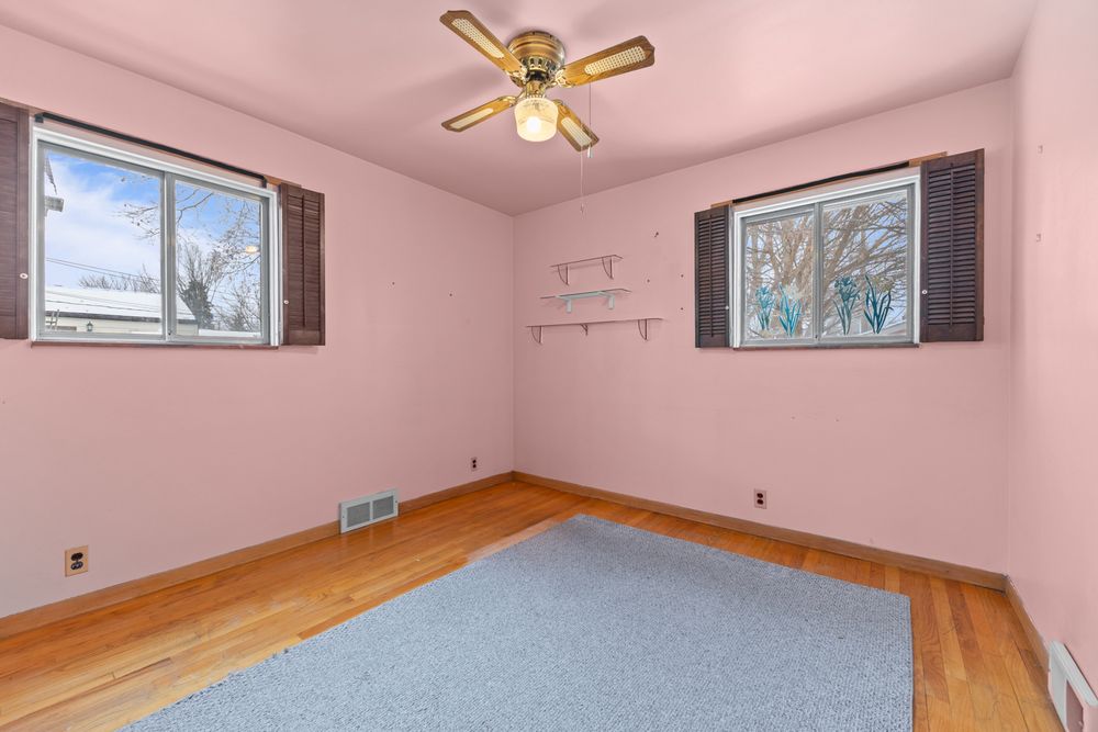 Bright empty bedroom with hardwood floors, ceiling fan, two windows, and light blue area rug.