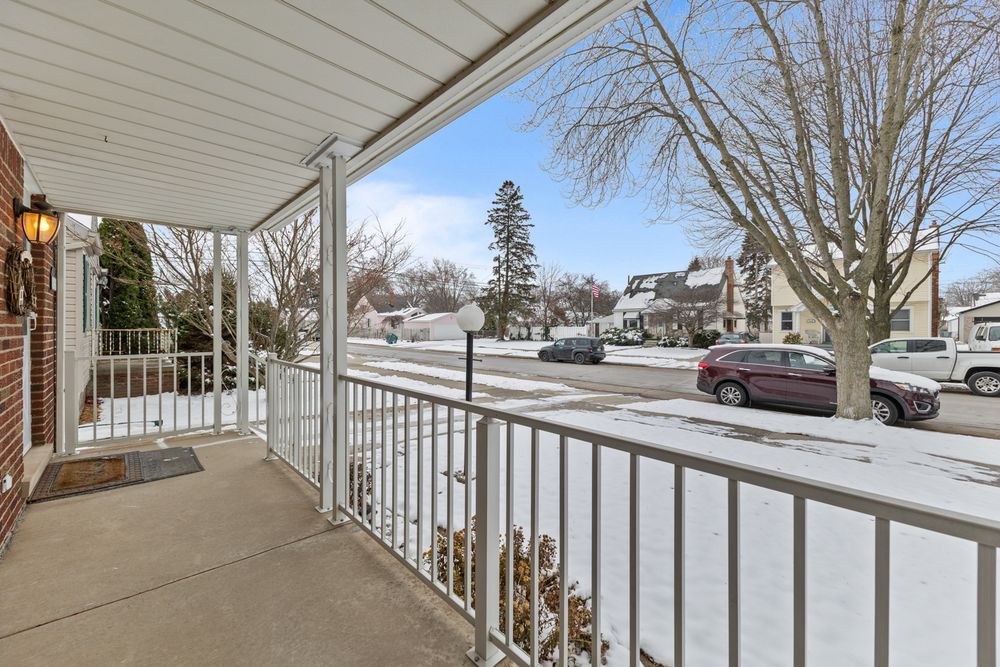 Snow-covered front porch with white railings overlooking a quiet residential street with parked cars and bare winter trees.