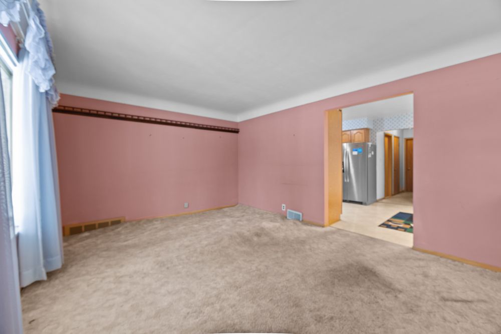 Empty room with pink walls, beige carpet, crown molding, and a doorway leading to a kitchen with stainless steel refrigerator.