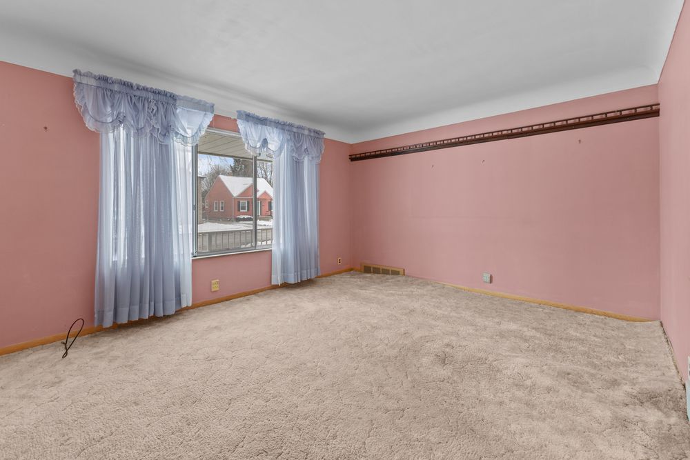 Empty bedroom with pink walls, beige carpet, and a window with light blue curtains overlooking a brick building.