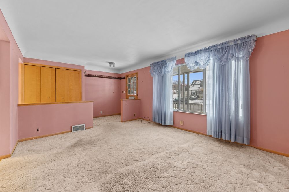 Spacious living room with pink walls, beige carpet, wood cabinetry, and a bright window with blue curtains.