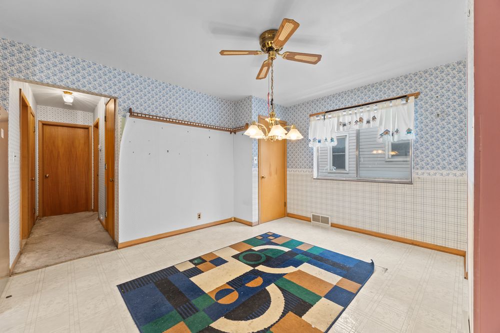 Vacant dining room with blue wallpaper, ceiling fan, chandelier, and colorful geometric area rug on beige carpet.