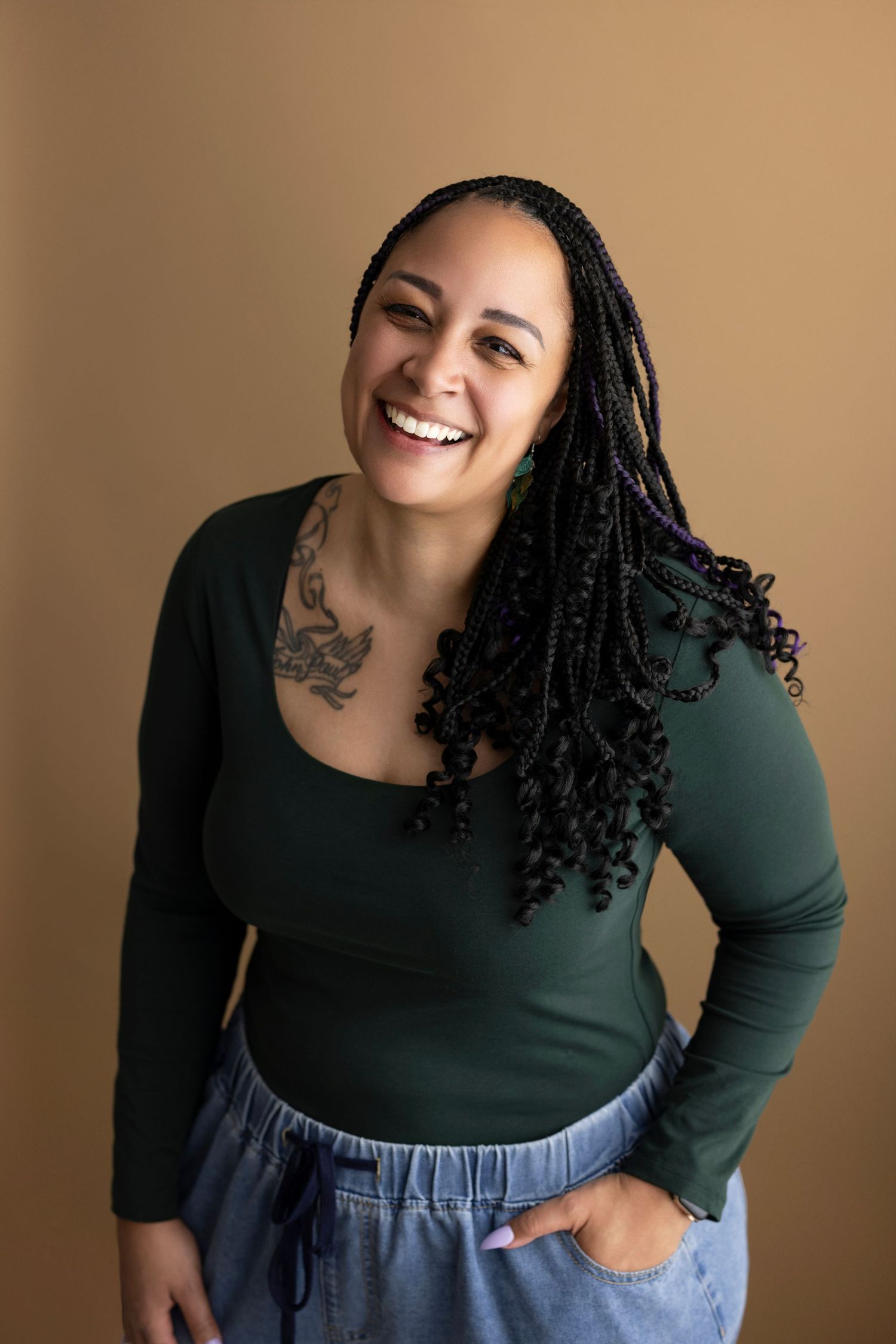 Smiling woman with long black braids, chest tattoo, green long-sleeve top, and jeans poses against a tan background.