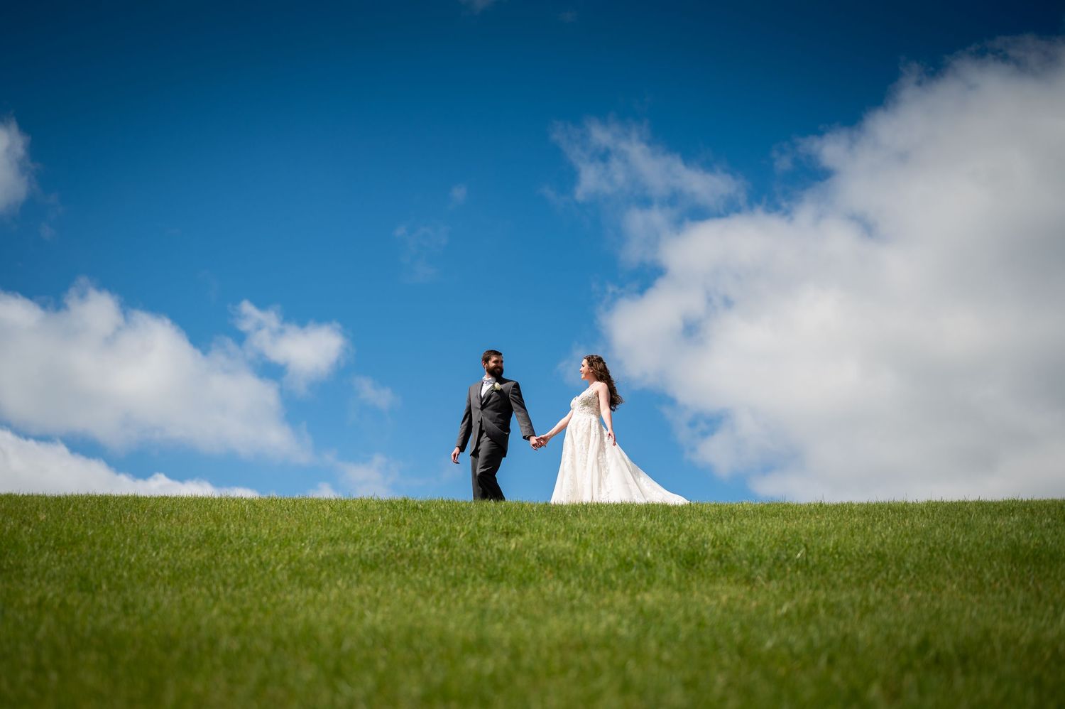 A bride and groom hold hands on a lush green hill under a bright blue sky with white clouds.