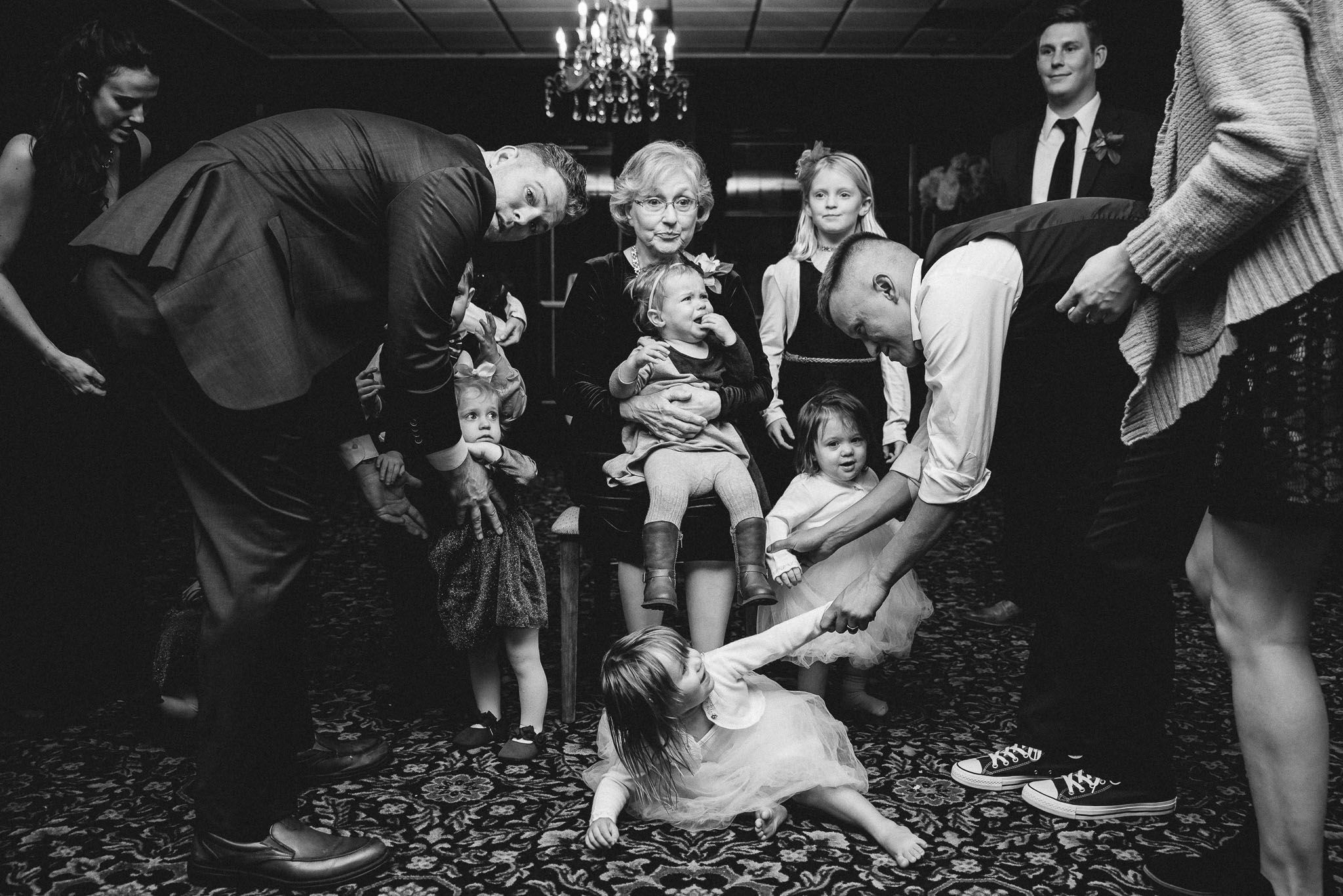 Black and white wedding reception photo of guests dancing with children on a patterned floor under a chandelier.